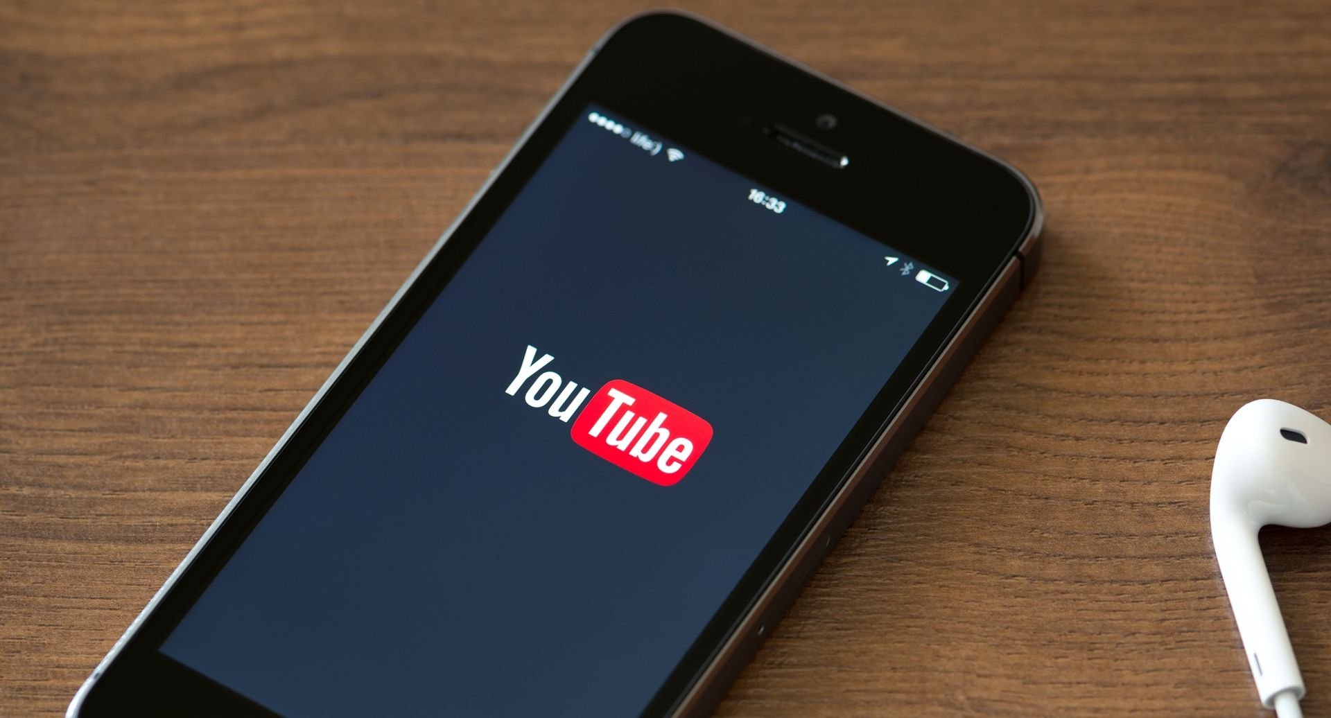 Um celular em cima de uma mesa e ao lado de fones de ouvido brancos, com a tela de abertura do YouTube ligada.