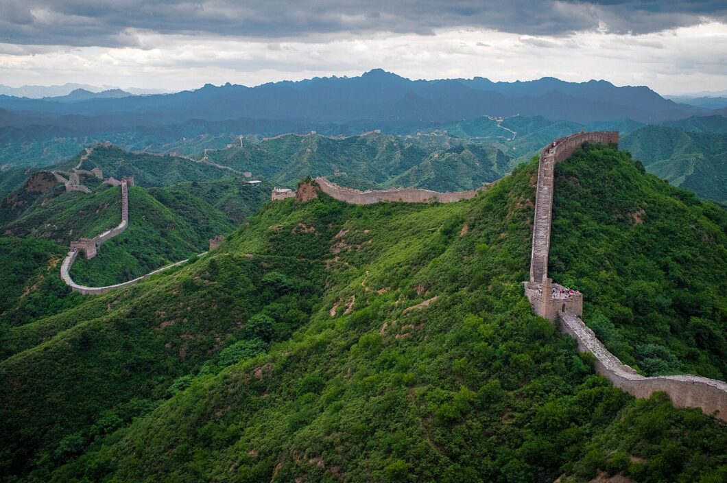 Muralha da China. Longa estrutura de muros com um corredor acima deles, que se estende por montanhas de vegetação alta.