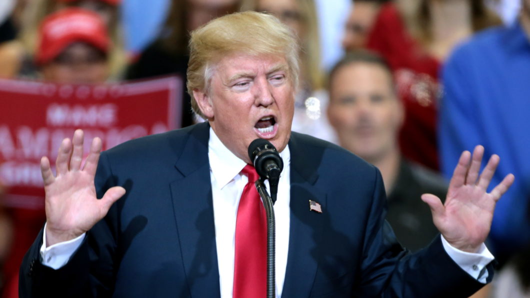 A imagem mostra Donald Trump de terno azul-escuro, camisa branca e gravata vermelha, falando em um microfone durante um evento público. Ele está com as duas mãos levantadas, com as palmas voltadas para frente, em um gesto expressivo. Um pequeno pin com a bandeira dos Estados Unidos está preso na lapela de seu paletó. Ao fundo, há várias pessoas desfocadas, muitas vestindo vermelho. O cenário indica que se trata de um evento de campanha eleitoral ou discurso público.