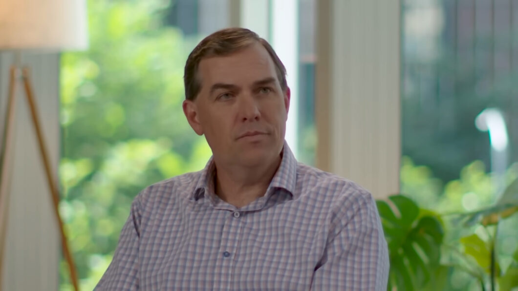 Matt Garman, CEO da AWS. Ele é um homem de meia-idade, veste camisa social xadrez em tons claros e olha levemente para o lado, com expressão séria. Ele está em um ambiente interno iluminado por luz natural, com plantas verdes ao fundo e uma luminária de pé à esquerda.