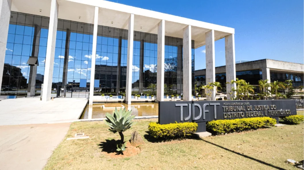 Esta imagem mostra o prédio do **Tribunal de Justiça do Distrito Federal e Territórios (TJDFT)**, que é o Tribunal de Justiça do Distrito Federal e Territórios no Brasil. A arquitetura apresenta elementos de design moderno, com grandes painéis de vidro e colunas verticais. O prédio parece ser cercado por uma paisagem bem cuidada, incluindo plantas e uma fonte de água na frente. O vidro refletivo oferece uma visão clara do céu e das nuvens, destacando o clima ensolarado.