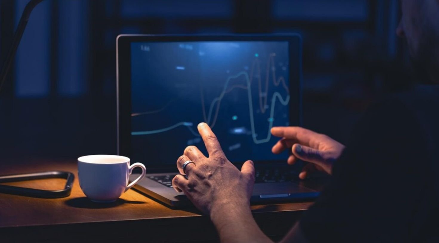 Um computador exibindo um gráfico variável de uma moeda, com uma pessoa em frente coberta por sombras e uma xícara de café na mesa.