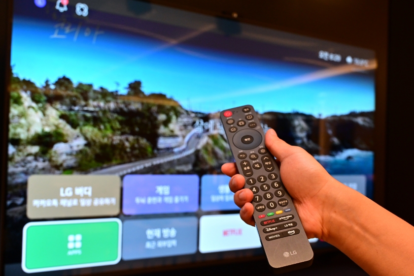 Uma mão segura um controle remoto preto da marca LG em primeiro plano, apontando-o para uma televisão ligada