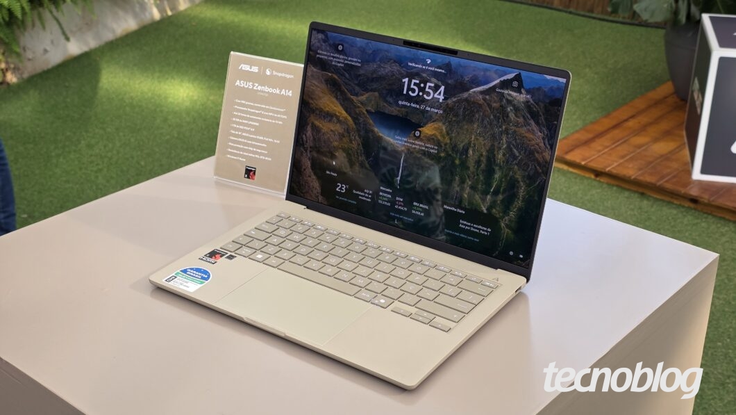 A imagem mostra um notebook ASUS Zenbook A14 exposto sobre uma mesa em um ambiente bem iluminado e decorado com grama sintética e plantas ao fundo. Ao lado do notebook, há um pequeno display informativo com o nome e algumas especificações do modelo, destacando o processador Snapdragon. A tela do notebook exibe o sistema Windows com o recurso de tela de bloqueio ativo, mostrando o horário (15:54), a data (quinta-feira, 21 de março) e informações sobre o clima e localização. O design do laptop é moderno e elegante, com acabamento metálico claro e teclado em tom semelhante. No canto inferior direito da imagem, aparece o logo do site Tecnoblog.
