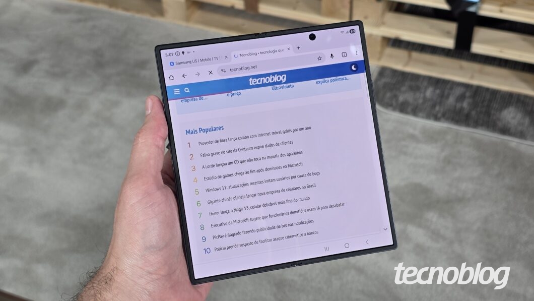Mão segurando smartphone dobrável aberto, exibindo a página do Tecnoblog