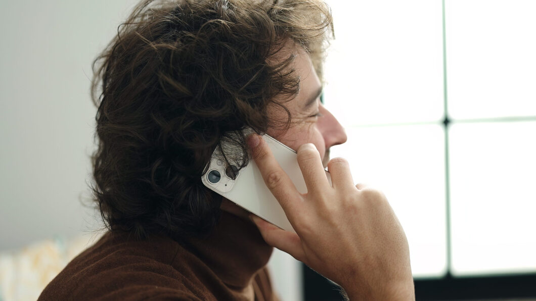 Imagem mostra uma pessoa sentada em um sofá falando ao telefone. O celular, segurado pela mão direita no ouvido, é um iPhone de cor branca.