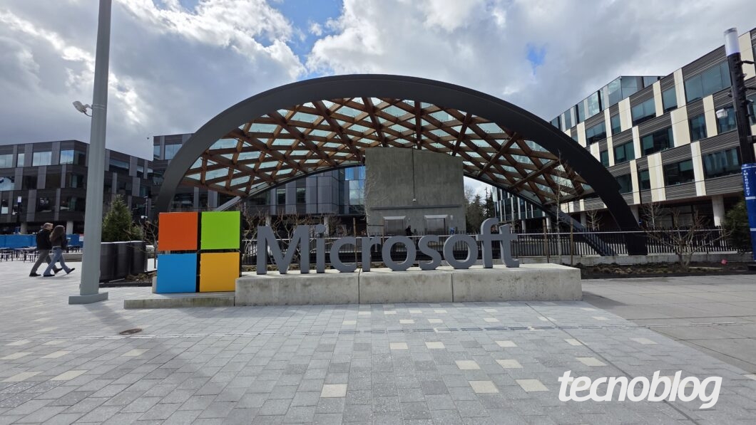 Imagem da entrada do campus da Microsoft em Redmond, nos Estados Unidos. Em destaque, o letreiro com o nome "Microsoft" ao lado do logo colorido da empresa. Ao fundo, há um edifício moderno com uma cobertura de madeira e vidro em arco. O céu está parcialmente nublado e algumas pessoas caminham pela calçada.