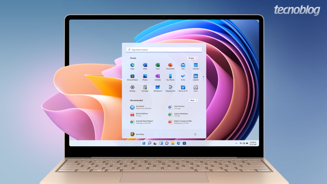 Um notebook Samsung com a tela exibindo a interface do Windows 11. O menu Iniciar está aberto ao centro, mostrando ícones de aplicativos e seções de "Recommended". O papel de parede do sistema operacional apresenta formas abstratas em tons de laranja, rosa, azul e roxo. No canto superior direito da imagem, há o logotipo "tecnoblog".
