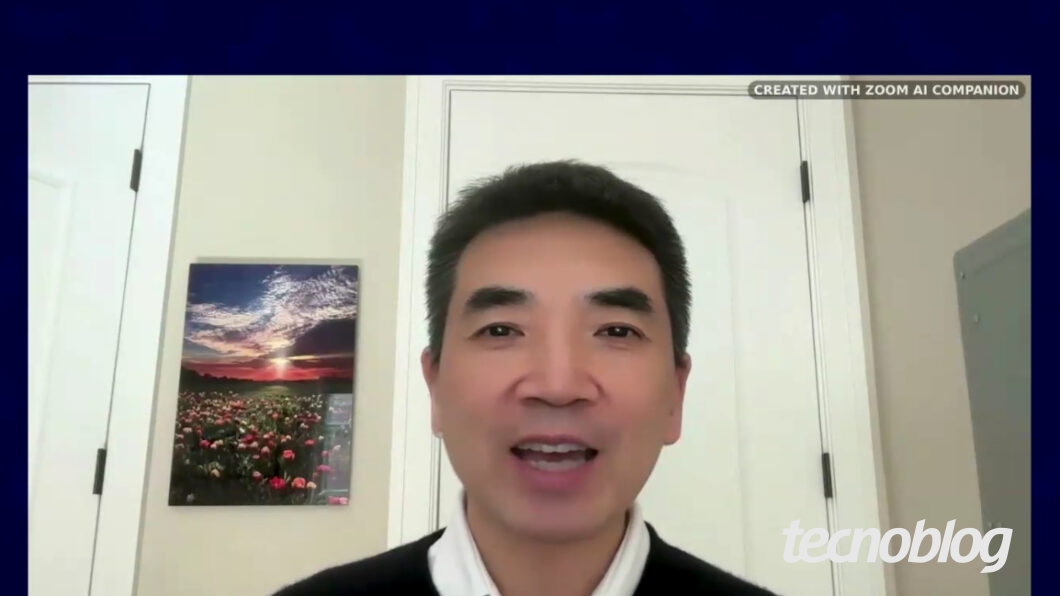 Captura de tela de uma videochamada do Zoom mostrando um homem falando em frente a uma porta branca. No canto superior direito, aparece a frase “CREATED WITH ZOOM AI COMPANION”. À esquerda, há um quadro com a imagem de um campo de flores e o pôr do sol.