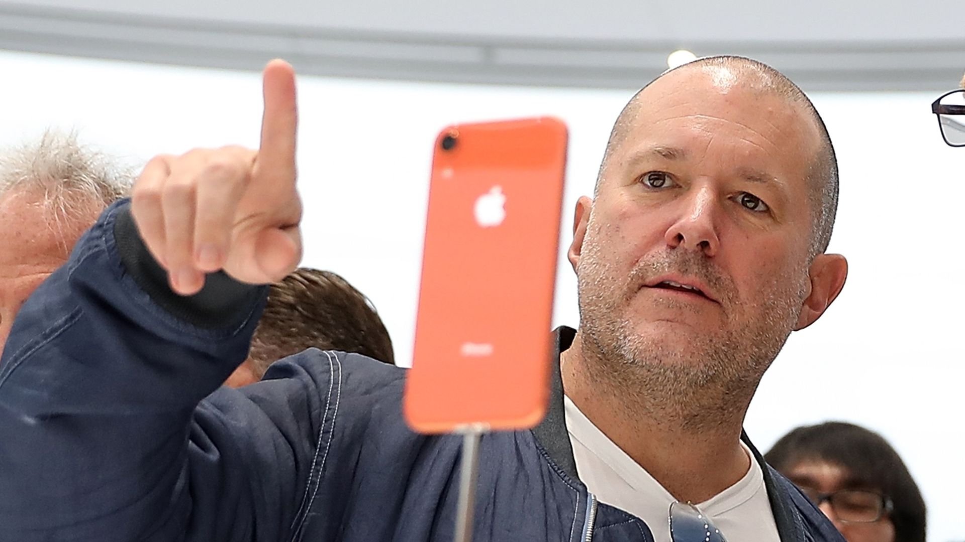 jony ive apontando para algo com o dedo indicado, atrás de um iPhone laranja suspenso por uma haste